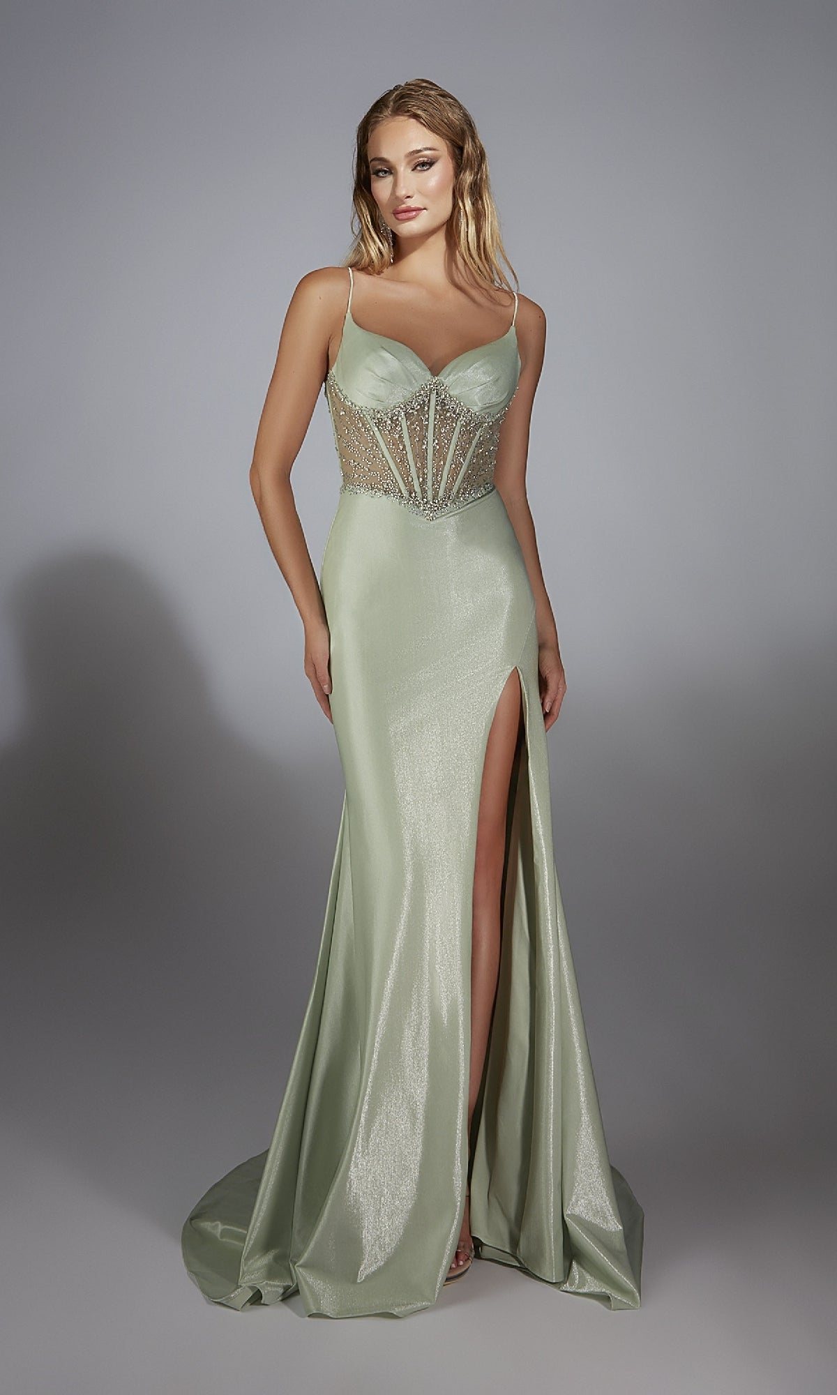 A woman models the Alyce 61916 long prom dress featuring a light green shimmer fabric, beaded corset bodice, spaghetti straps, and high thigh slit, standing against a plain gray background.